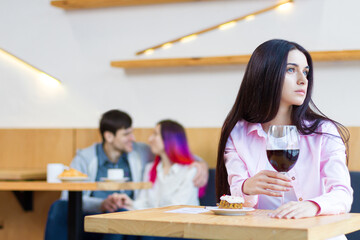 Lonely sad girl is sitting alone at table in cafe, restaurant. Young woman without relationships is spending time with herself. Loving couple is on date on background. Loneliness concept.
