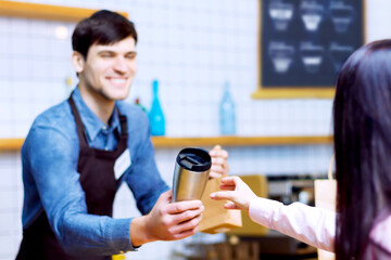 Barista man in brown apron is giving packed order to woman. Guy made coffee to go in metal reusable eco thermo cup for client. Takeaway work of restaurants, cafes. Customer service concept.