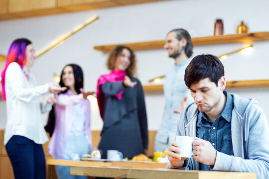 Closeup portrait of sad man, outcast is sitting alone at table in cafe. Teenagers, students are mocking, scoffing, pointing at guy. Influence of society on mental health. Bullying concept.