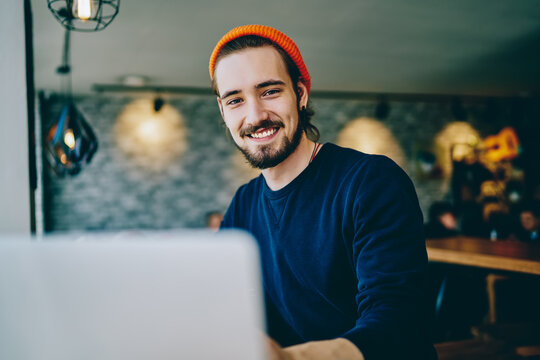 Portrait of cheerful caucasian man in trendy bennie satisfied with online language courses using netbook for studying, happy bearded hipster looking at camera satisfied with wifi for remote work