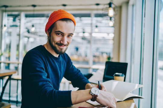 Portrait of cheerful caucasian hipster guy checking time for learning in cafe interior, happy male freelancer looking at camera showing wristwatch using time management for complete remote job.