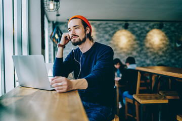 Pensive hipster guy sitting on publicity area with laptop computer and listening audio book about...
