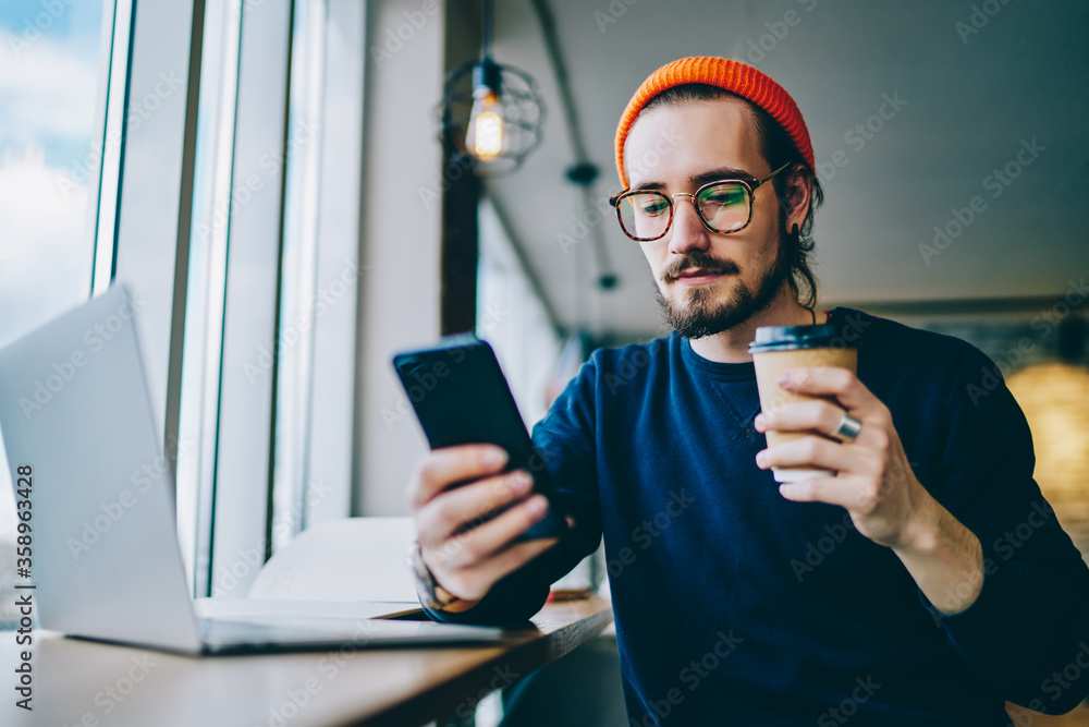 Poster male hipster in eyeglasses searching media files on cellular phone for sharing with laptop via bluet