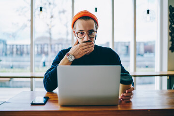 Emotional hipster guy in spectacles read amazing news on laptop computer while working remotely,...