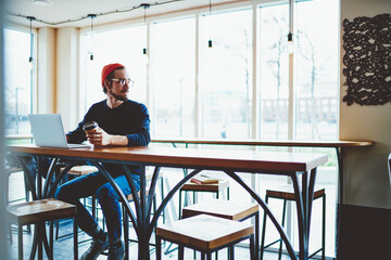 Fototapeta premium Pondering hipster guy thoughtful looking away and thinking about information database on laptop computer during distance job in coffee shop, Caucasian freelancer have freelance work on netbook