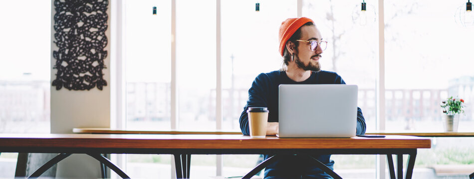 Pondering Hipster Guy Thoughtful Looking Away And Thinking About Information Database On Laptop Computer During Distance Job In Coffee Shop, Front View Of Caucasian Freelancer Sitting Indoors