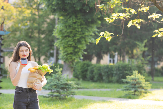 Happy girl using a smart phone voice recognition audio ai message speech function on line walking on a park background and carrying bag with food