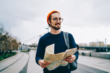 Cheerful caucasian hipster guy in trendy spectacles satisfied with active vacation having trip , smiling male traveler strolling with map having city tour excited with urban setting on weekends.