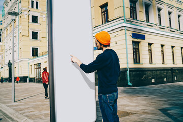 Back view of caucasian male traveler searching information on touristic billboard pointing on white copy space area, hipster guy showing content on blank frame placeholder standing outdoors on street.