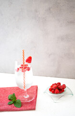 Refreshing raspberry lemonade in a glass with a cocktail straw is on a red napkin and on a white table. Nearby are mint leaves and a bowl of fresh raspberries. Vertically