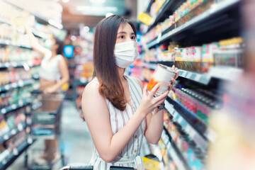 Asian woman wearing face mask push shopping cart in suppermarket . Girl choosing, looking somethings to buy at shelf during coronavirus crisis or covid19 .happy lifestyle or shopaholic concept.