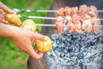 Hands of man prepares barbecue meat with potatoes on skewer by grill on fire outdoors. Concept of lifestyle rustic food preparation