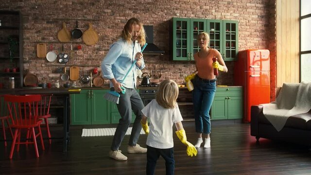 Young parents and their kid in yellow rubber gloves are smiling and dancing with detergent and broom at kitchen with modern interior. Slow motion