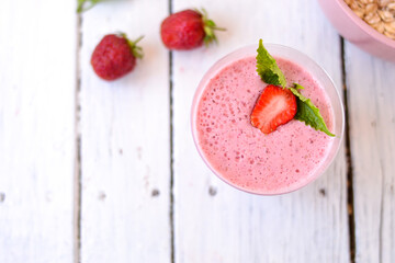 Cocktail with strawberries and milk in a glass decorated with mint and strawberries. Nearby is a pink cup with Hercules flakes, and loose strawberries on a white wooden background. 