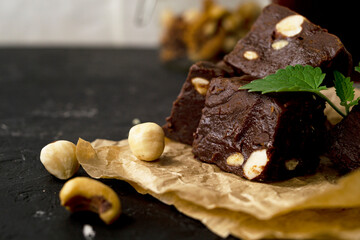 Fondant on craft crumpled paper with nuts and chocolate cubes decorated with green mint leaves. In the background a jar of nuts, blurred background. Sweet composition on a black textural background. 