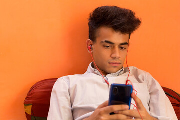 A Young Man listening to music on his cellphone while lying down over a pillow on a bed.