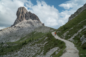 Hiker with orange backpack on mountain trail passing between two mountains in Dolomites, Italy.