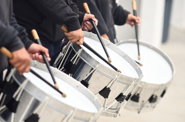 members of a war band wearing black and loading their silver drums with thick drumsticks covered in black ribbon, playing rhythms in unison
