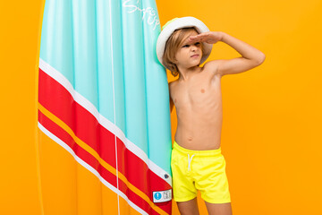 Handsome boy in swimming trunks holds a rubber mattress, smiles and gesticulates isolated on orange...