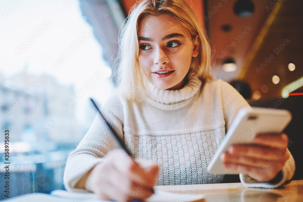Poster clever female student doing homework task in cafe interior holding smartphone for networking,creativ