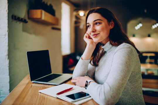 Dreamy female teen student thinking about vacation while preparing homework in cafe interior, positive young woman lost in her thoughts while working on freelance at desktop in coworking space .