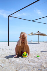 Lovely funny little boy sit at sandy sea beach, cover from sun, play with toy plastic concrete...