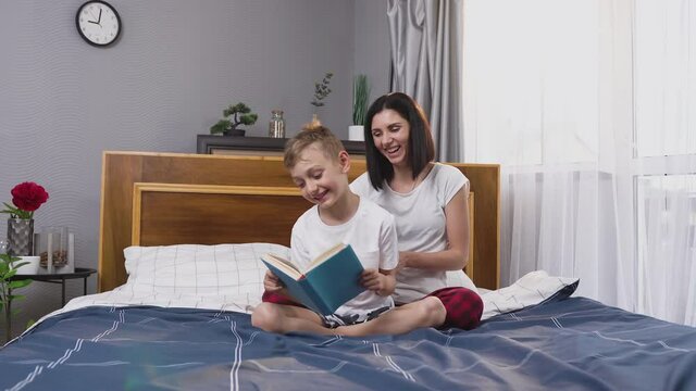 Attractive happy 8-aged boy reading book on bedroom bed while his smiling loving mother tickling him