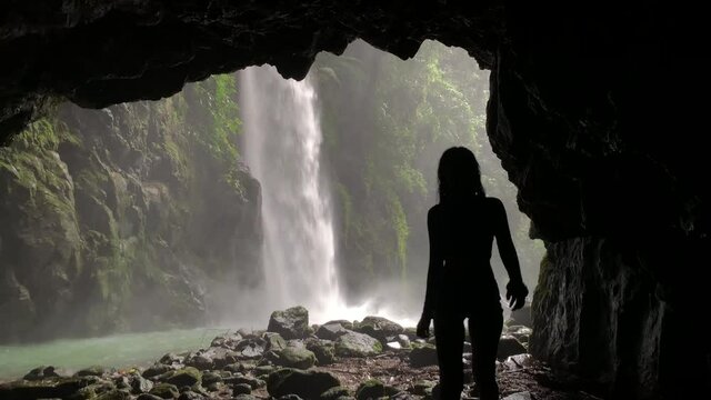 Silhouette of a woman walking inside the cave in a rainforest waterfall.