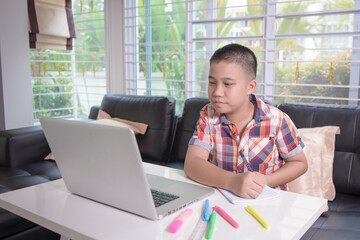 Asian male student is sitting at home and doing his homework on a laptop. He is reading the screen with a thoughtful intentionally. Home learning concepts.