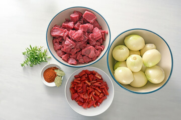 Ingredients for stew or goulash with diced beef, onions, bell pepper, herbs and spices from above on a white painted table, cooking at home during the corona virus pandemic