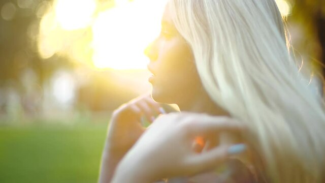 Close-up profile shot of a stunning young blonde in lightweight dress fluffy long hair against spring park in sunny weather. Beautiful girl with bright manicure enjoys warm spring in cozy city park.