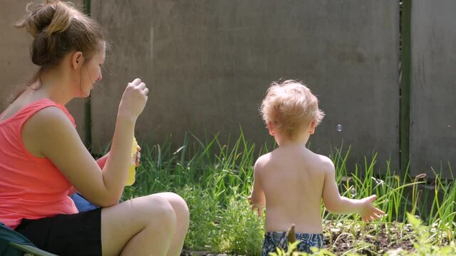Mother with son blow bubbles soap young woman 30 years old and naked toddler boy 2 years old play in backyard slow motion