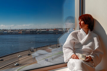 Beautiful middle age woman drinking tea sitting by the window. Middle-aged woman rests at panoramic window. River and city landscape, sunny day with clear blue sky