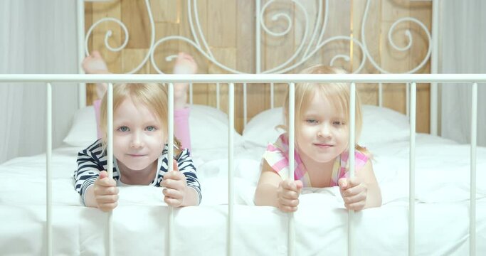 Portrait Of Smiling Cute Sisters Looking At Camera Lying On Bed And Dangling Feet Together At Home.