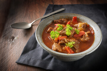 Hot goulash or stew, cooked from beef, onions and red bell pepper with parsley garnish in a bowl with spoon and a dark napkin on a rustic wooden table