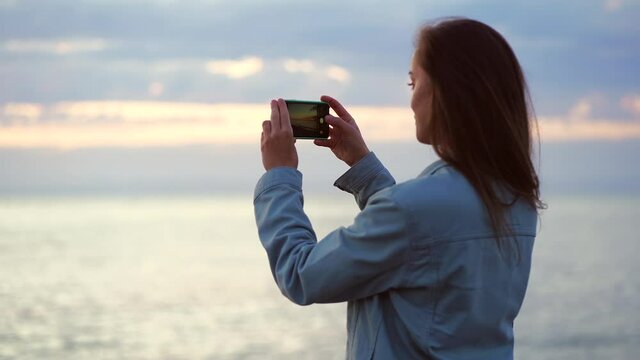 Young casual woman traveler taking picture of the sea at sunset on the mobile phone camera 