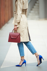 Fashionable young woman in cropped jeans, blue blouse, beige trench and two handbag .She is wearing blue high heel shoes. Street style.