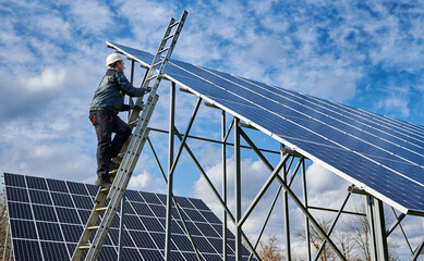 Full length of man electrician in safety helmet climbing the ladder to get to the top of photovoltaic solar module. Technician mounting solar photovoltaic panel system under beautiful cloudy sky.