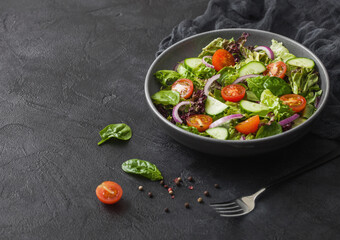 Fresh vegetables salad with lettuce and tomatoes, red onion and spinach in black bowl on dark background with dinner fork