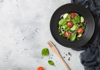 Fresh healthy vegetarian vegetables salad with lettuce and tomatoes, red onion and spinach in black bowl on dark background with spatula fork and black kitchen cloth.