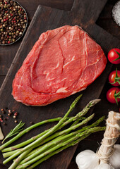 Slice of raw fresh beef braising steak on chopping board with garlic, asparagus and tomatoes with salt and pepper on wooden background.