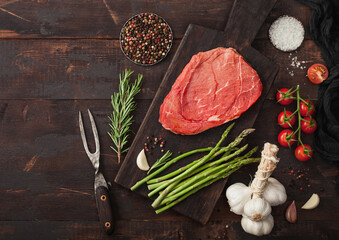 Fresh raw slice of beef braising steak on chopping board with asparagus, garlic and tomatoes with salt and pepper on wooden background.