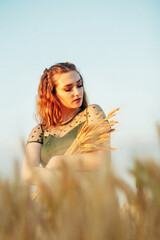 Beautiful woman in green dress on field with sheaf of wheat in hand at sunset light, girl enjoying summer nature landscape, harvest