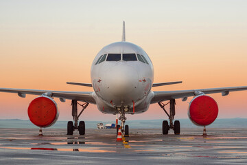 Front view of white airplane. Jet commercial aircraft on airport apron, morning sunrise orange red sky, rain puddles. Modern technology in fast transportation, private business travel, charter flights