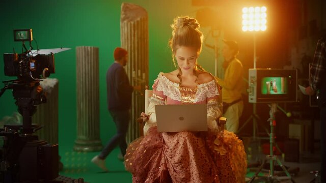  Beautiful Smiling Actress Wearing Renaissance Dress, Sitting on a Chair Using Laptop Computer with Green Screen in the Background. On Film Studio Period Costume Drama Film Set