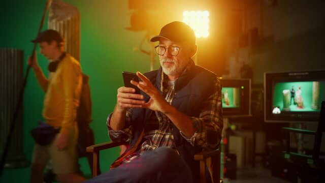 Prominent Successful Senior Director Sitting in a Chair on a Break Using Smartphone. On the Studio Film Set with High-End Equipment Professional Crew Shooting High Budget Movie