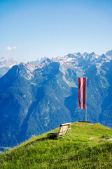 Austrian Flag waving in the wind next to a wooden bench at a grass covered Mountain peak with clear blue sky and the austrian alps in the background at a summer day