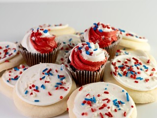 White sugar cookies with red, white and blue sprinkles with 3 mini chocolate cupcakes with red and white swirled frosting and star sprinkles sitting on top for the 4th of July.