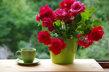 A bouquet of red roses in a green vase and a cup of tea on the windowsill.