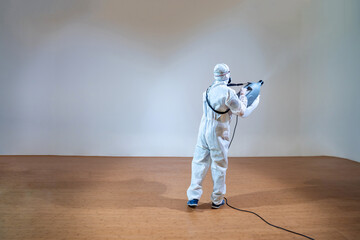 Professional technical man in prevention suit sprays sterilising solution by electrical spray machine on wood floor and white background with studio light.